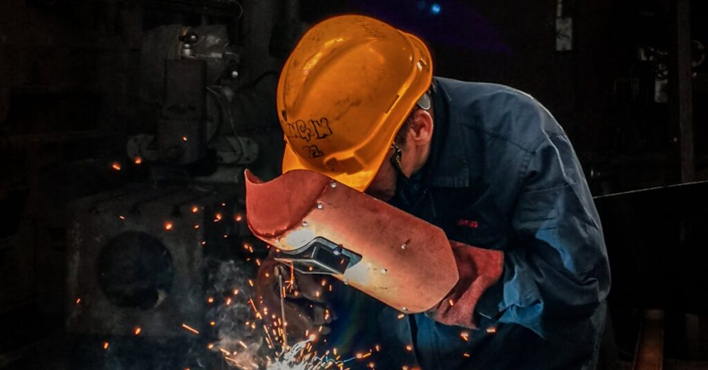 A welder wearing protective gear works on metal in an industrial workshop, surrounded by sparks.